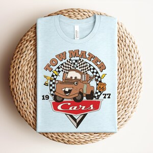 Puede incluir: Una camiseta azul claro con un gr&aacute;fico del personaje Tow Mater de la pel&iacute;cula Cars. El dise&ntilde;o incluye una bandera a cuadros, un trofeo dorado, una se&ntilde;al de la Ruta 66, el texto Tow Mater, 1977 y el logotipo de Cars en rojo y blanco.