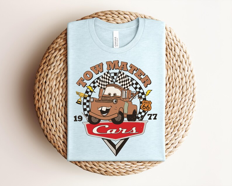 Puede incluir: Una camiseta azul claro con un gr&aacute;fico del personaje Tow Mater de la pel&iacute;cula Cars. El dise&ntilde;o incluye una bandera a cuadros, un trofeo dorado, una se&ntilde;al de la Ruta 66, el texto Tow Mater, 1977 y el logotipo de Cars en rojo y blanco.