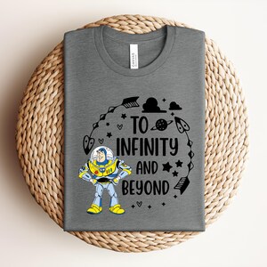 Peut inclure: Un t-shirt gris avec un motif color&eacute; de Buzz l'&Eacute;clair et le texte "TO INFINITY AND BEYOND" en noir. Le motif comprend des &eacute;toiles, des fl&egrave;ches et une plan&egrave;te. Le t-shirt est pli&eacute; sur une surface tiss&eacute;e.
