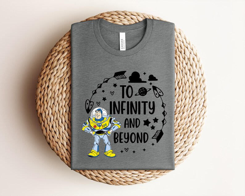 Peut inclure: Un t-shirt gris avec un motif color&eacute; de Buzz l'&Eacute;clair et le texte "TO INFINITY AND BEYOND" en noir. Le motif comprend des &eacute;toiles, des fl&egrave;ches et une plan&egrave;te. Le t-shirt est pli&eacute; sur une surface tiss&eacute;e.