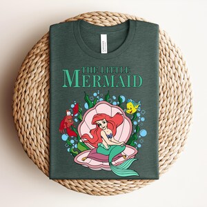 May include: A dark forest green t-shirt featuring a graphic of The Little Mermaid with Ariel sitting in a pink seashell, accompanied by Sebastian and Flounder, surrounded by green seaweed and blue bubbles, with the text The Little Mermaid above.
