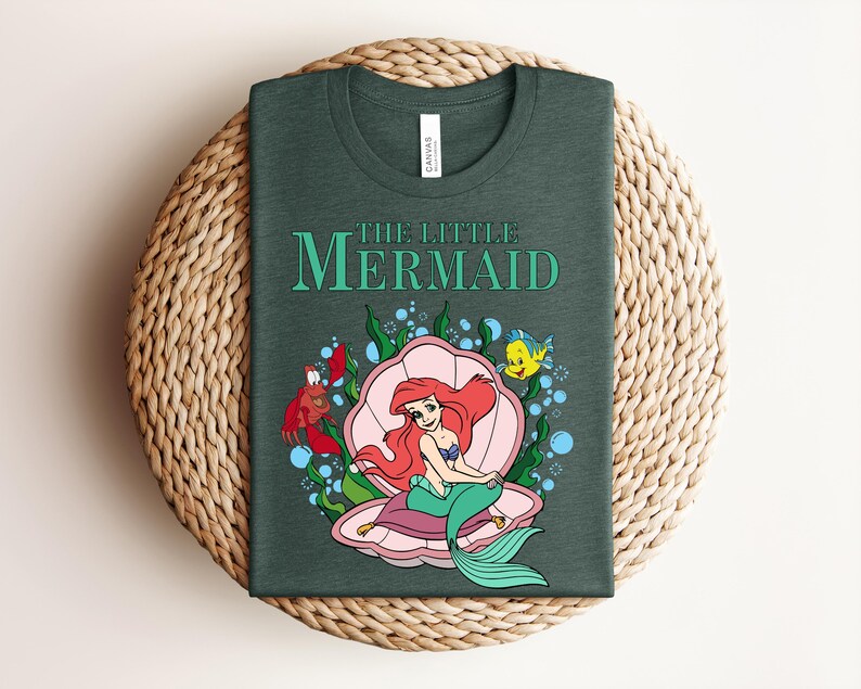 May include: A dark forest green t-shirt featuring a graphic of The Little Mermaid with Ariel sitting in a pink seashell, accompanied by Sebastian and Flounder, surrounded by green seaweed and blue bubbles, with the text The Little Mermaid above.