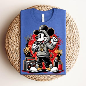 May include: Blue t-shirt featuring a cartoon Mickey Mouse character in a hip-hop style. Mickey is wearing a hoodie, holding a microphone, and a gold chain. Graffiti art and a boombox are also in the design.