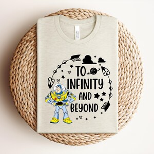 Peut inclure: Un t-shirt beige clair avec un motif de Buzz l'&Eacute;clair et l'inscription "TO INFINITY AND BEYOND" en noir. Le motif comprend des &eacute;toiles, des plan&egrave;tes et des &eacute;l&eacute;ments sur le th&egrave;me de l'espace. Le t-shirt est pr&eacute;sent&eacute; sur un tapis circulaire tiss&eacute;.