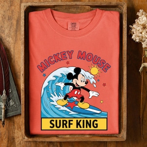 May include: Coral-colored t-shirt featuring Mickey Mouse surfing a wave. The graphic includes the words "Mickey Mouse" and "Surf King." The design has a cartoon sun, stars, and a blue surfboard. The shirt is folded in a wooden tray.