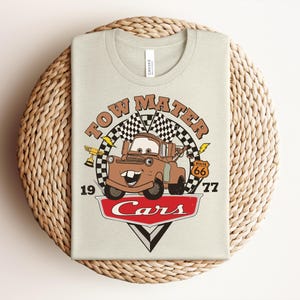 Puede incluir: Una camiseta de color beige claro con un gr&aacute;fico del personaje Tow Mater de la pel&iacute;cula Cars. El dise&ntilde;o incluye una bandera a cuadros, un trofeo dorado, una se&ntilde;al de la Ruta 66, el texto Tow Mater, 19, 77 y el logo rojo de Cars.