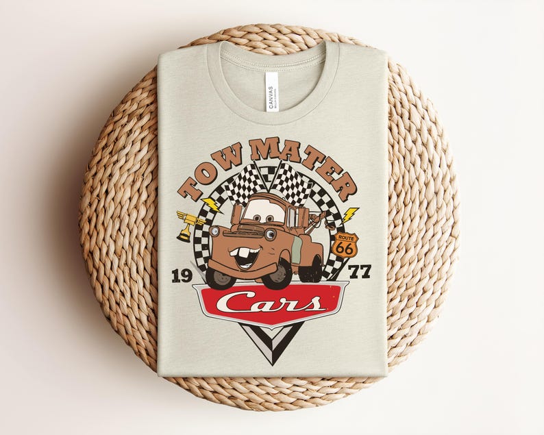 Puede incluir: Una camiseta de color beige claro con un gr&aacute;fico del personaje Tow Mater de la pel&iacute;cula Cars. El dise&ntilde;o incluye una bandera a cuadros, un trofeo dorado, una se&ntilde;al de la Ruta 66, el texto Tow Mater, 19, 77 y el logo rojo de Cars.