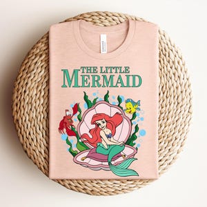 May include: A light peach-colored t-shirt featuring a graphic of Ariel from The Little Mermaid sitting in a pink seashell. The design includes green seaweed, a red crab, and a yellow fish, with the text THE LITTLE MERMAID printed in green.