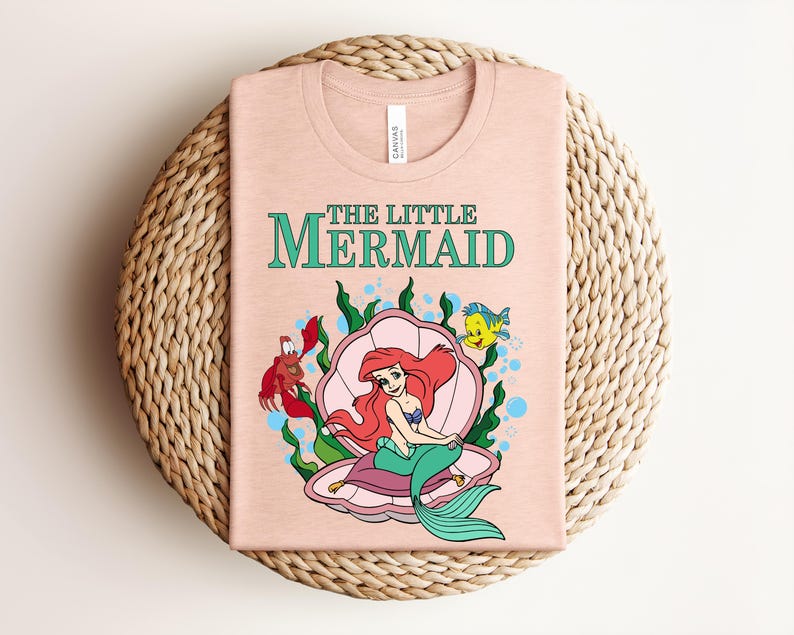 May include: A light peach-colored t-shirt featuring a graphic of Ariel from The Little Mermaid sitting in a pink seashell. The design includes green seaweed, a red crab, and a yellow fish, with the text THE LITTLE MERMAID printed in green.