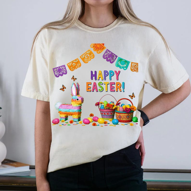 K&ouml;nnte beinhalten: Cremefarbenes T-Shirt mit einem farbenfrohen "Frohe Ostern!"-Motiv. Das Design umfasst einen Bunny im Pi&ntilde;ata-Stil, Osterk&ouml;rbe, Eier und dekorative Banner. Schmetterlinge verleihen dem festlichen Thema eine verspielte Note.