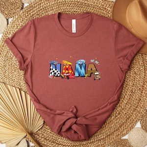 K&ouml;nnte beinhalten: Ein staubrosafarbenes T-Shirt mit dem Wort "NANA" in bunten Buchstaben im Autothema. Die Buchstaben sind mit Rennwagen und Zielflaggen verziert. Das Shirt ist aus weichem Material.