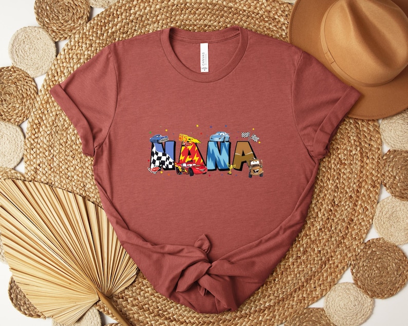 K&ouml;nnte beinhalten: Ein staubrosafarbenes T-Shirt mit dem Wort "NANA" in bunten Buchstaben im Autothema. Die Buchstaben sind mit Rennwagen und Zielflaggen verziert. Das Shirt ist aus weichem Material.