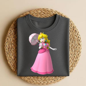 May include: A dark gray t-shirt featuring a graphic print of Princess Peach from the Super Mario series. The character is depicted with blonde hair, a pink dress, and a light pink parasol. The design is centered on the front of the shirt.