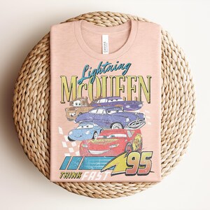 Puede incluir: Una camiseta color melocot&oacute;n con un gr&aacute;fico de Rayo McQueen y otros personajes de Cars. El dise&ntilde;o incluye el texto "Lightning McQueen" y "95", con la frase "Think Fast" debajo. La camiseta est&aacute; doblada sobre una superficie circular tejida.