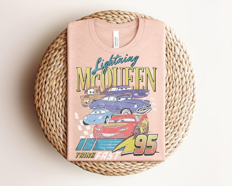 Puede incluir: Una camiseta color melocot&oacute;n con un gr&aacute;fico de Rayo McQueen y otros personajes de Cars. El dise&ntilde;o incluye el texto "Lightning McQueen" y "95", con la frase "Think Fast" debajo. La camiseta est&aacute; doblada sobre una superficie circular tejida.