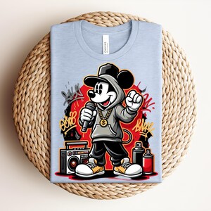 May include: Light blue t-shirt featuring a cartoon Mickey Mouse character in a hip-hop style. Mickey is wearing a gray hoodie, black pants, and yellow shoes, holding a microphone. The design includes graffiti art, a boombox, and spray paint cans.