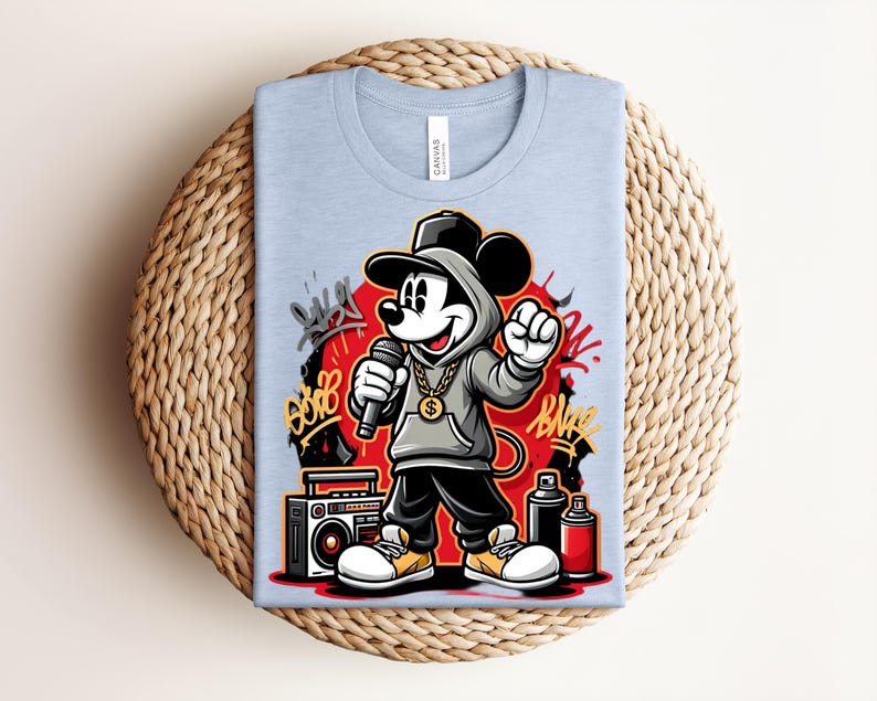 May include: Light blue t-shirt featuring a cartoon Mickey Mouse character in a hip-hop style. Mickey is wearing a gray hoodie, black pants, and yellow shoes, holding a microphone. The design includes graffiti art, a boombox, and spray paint cans.