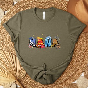 K&ouml;nnte beinhalten: Olivgr&uuml;nes T-Shirt mit dem Wort "NANA" in bunten Buchstaben, die jeweils als Rennwagen gestaltet sind. Die Autos sind in verschiedenen Farben und Mustern gehalten, darunter karierte Flaggen und Flammen. Das Shirt besteht aus weichem Material.