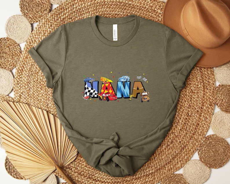 K&ouml;nnte beinhalten: Olivgr&uuml;nes T-Shirt mit dem Wort "NANA" in bunten Buchstaben, die jeweils als Rennwagen gestaltet sind. Die Autos sind in verschiedenen Farben und Mustern gehalten, darunter karierte Flaggen und Flammen. Das Shirt besteht aus weichem Material.