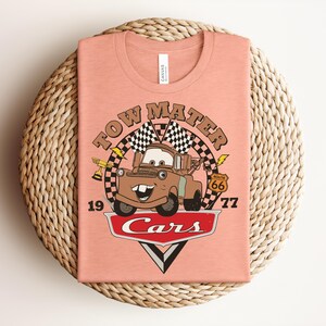 Puede incluir: Una camiseta color melocot&oacute;n con un gr&aacute;fico del personaje Tow Mater de la pel&iacute;cula Cars. El dise&ntilde;o incluye el texto "TOW MATER", "1977", "ROUTE 66" y "Cars" junto con banderas a cuadros, un trofeo y detalles de rayos.