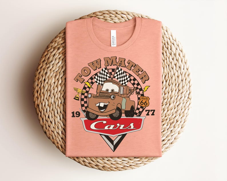 Puede incluir: Una camiseta color melocot&oacute;n con un gr&aacute;fico del personaje Tow Mater de la pel&iacute;cula Cars. El dise&ntilde;o incluye el texto "TOW MATER", "1977", "ROUTE 66" y "Cars" junto con banderas a cuadros, un trofeo y detalles de rayos.