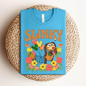 K&ouml;nnte beinhalten: Ein hellblaues T-Shirt mit dem Wort "SLINKY" in orangefarbenen Buchstaben. Das Design zeigt Slinky Dog, Ballons, Blumen und eine Biene. Das T-Shirt liegt gefaltet auf einer runden, gewebten Unterlage.