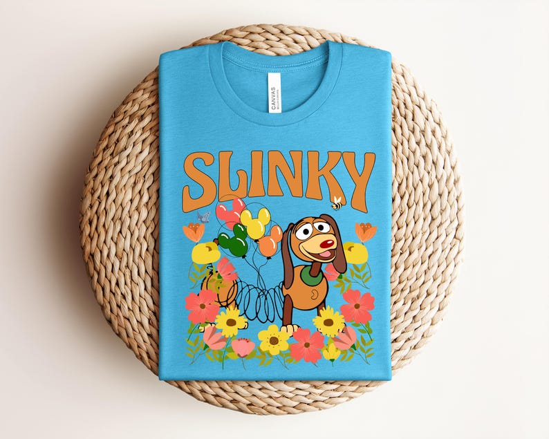 K&ouml;nnte beinhalten: Ein hellblaues T-Shirt mit dem Wort "SLINKY" in orangefarbenen Buchstaben. Das Design zeigt Slinky Dog, Ballons, Blumen und eine Biene. Das T-Shirt liegt gefaltet auf einer runden, gewebten Unterlage.