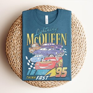 Puede incluir: Camiseta de color turquesa con un dise&ntilde;o de Rayo McQueen y otros personajes de Cars. El dise&ntilde;o incluye el texto "Lightning McQueen", "95" y "Think Fast" en un estilo vintage. La camiseta est&aacute; doblada sobre una estera tejida.