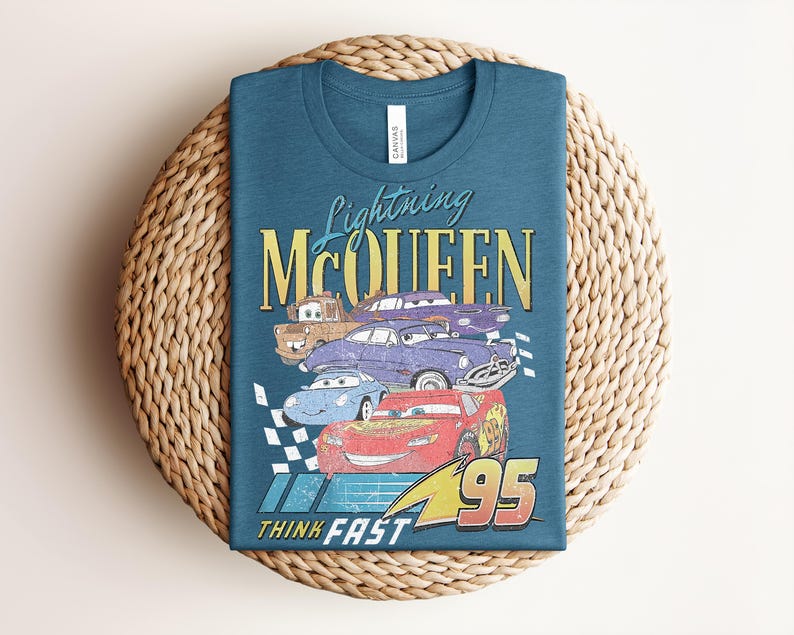Puede incluir: Camiseta de color turquesa con un dise&ntilde;o de Rayo McQueen y otros personajes de Cars. El dise&ntilde;o incluye el texto "Lightning McQueen", "95" y "Think Fast" en un estilo vintage. La camiseta est&aacute; doblada sobre una estera tejida.
