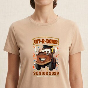 Op de afbeelding: Beige T-shirt met een cartoonafbeelding van Takel uit de film Cars, met een afstudeerhoed. De tekst "GIT-R-DONE!" staat boven de afbeelding en "SENIOR 2026" eronder. Het shirt heeft een ronde hals.