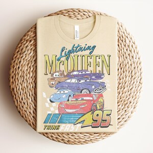 Puede incluir: Camiseta color beige con un dise&ntilde;o de Rayo McQueen y otros personajes de Cars. El dise&ntilde;o incluye el texto "Lightning McQueen" y "95", con la frase "Think Fast" debajo. La camiseta est&aacute; doblada sobre una superficie circular tejida.