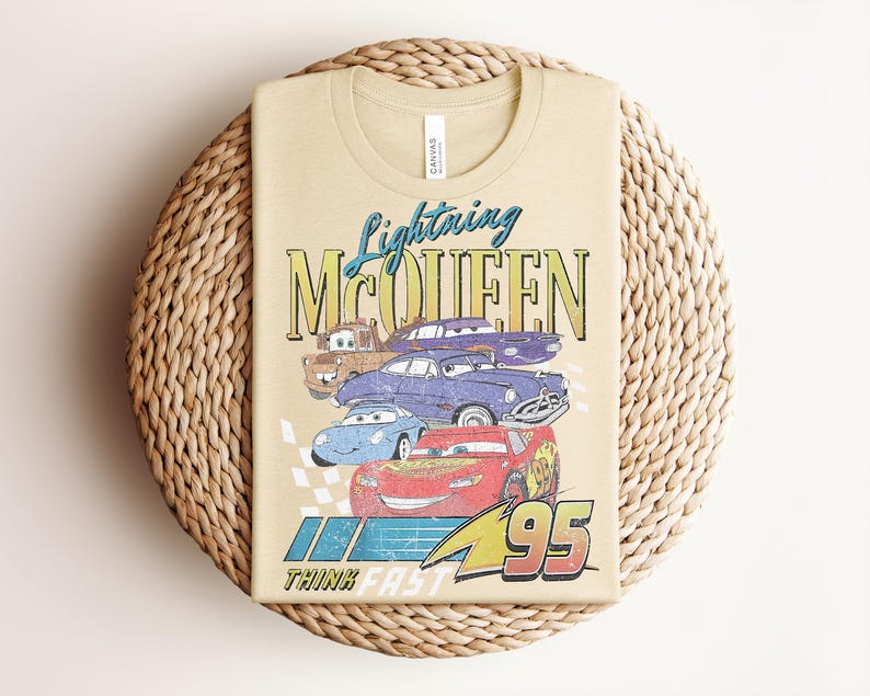 Puede incluir: Camiseta color beige con un dise&ntilde;o de Rayo McQueen y otros personajes de Cars. El dise&ntilde;o incluye el texto "Lightning McQueen" y "95", con la frase "Think Fast" debajo. La camiseta est&aacute; doblada sobre una superficie circular tejida.