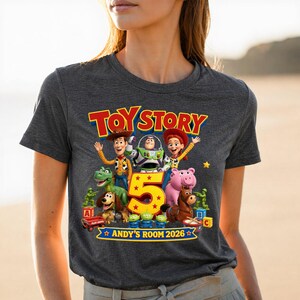 May include: A dark heather gray t-shirt featuring a colorful Toy Story graphic with characters including Woody, Buzz Lightyear, Jessie, Rex, Slinky Dog, Hamm, and Bullseye. The design includes a large yellow number 5 and the text ANDY'S ROOM 2026.