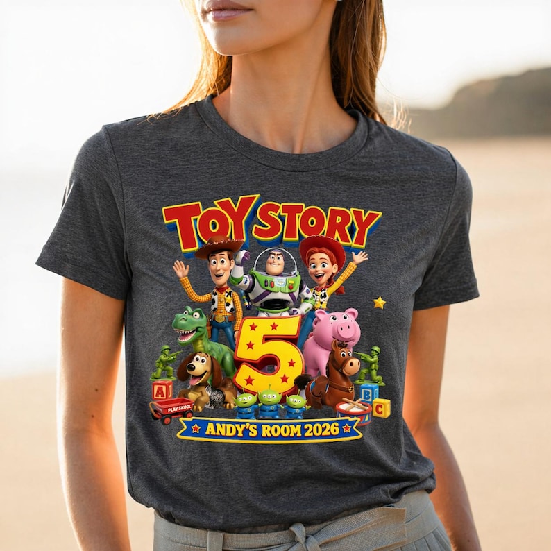 May include: A dark heather gray t-shirt featuring a colorful Toy Story graphic with characters including Woody, Buzz Lightyear, Jessie, Rex, Slinky Dog, Hamm, and Bullseye. The design includes a large yellow number 5 and the text ANDY'S ROOM 2026.