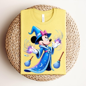 May include: Yellow t-shirt featuring a cartoon illustration of a mouse dressed as a wizard. The mouse is wearing a blue wizard hat with a pink bow and a blue robe. The design includes magic effects and two small brooms.