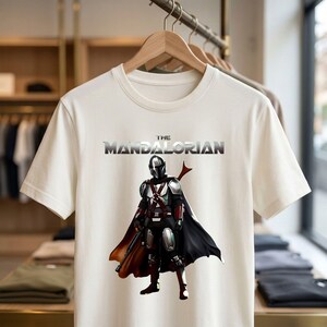 Op de afbeelding: Cr&egrave;mekleurig T-shirt met een afbeelding van The Mandalorian. Het ontwerp toont het personage in volledige uitrusting met een zwarte cape en een rood wapen. De tekst "THE MANDALORIAN" staat boven het personage in metallic zilver.