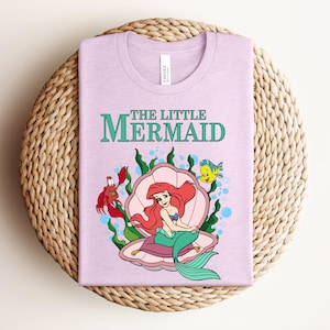 May include: A light pink t-shirt featuring a graphic of The Little Mermaid with Ariel sitting in a pink seashell, accompanied by Sebastian the crab and Flounder the fish, surrounded by green seaweed and blue bubbles on a light pink fabric.