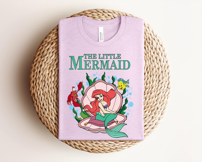 May include: A light pink t-shirt featuring a graphic of The Little Mermaid with Ariel sitting in a pink seashell, accompanied by Sebastian the crab and Flounder the fish, surrounded by green seaweed and blue bubbles on a light pink fabric.