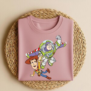 May include: A light pink crew neck sweatshirt featuring a colorful graphic of Buzz Lightyear and Woody from Toy Story. Buzz is in flight, and Woody is reaching out. The sweatshirt is folded on a woven mat.