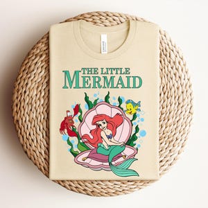 May include: A light beige t-shirt featuring a graphic of The Little Mermaid with Ariel sitting in a pink seashell, accompanied by a red crab and a yellow fish, surrounded by green seaweed and blue bubbles, with the text The Little Mermaid above.