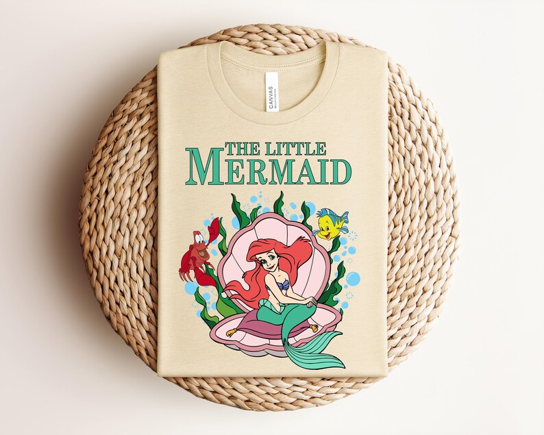 May include: A light beige t-shirt featuring a graphic of The Little Mermaid with Ariel sitting in a pink seashell, accompanied by a red crab and a yellow fish, surrounded by green seaweed and blue bubbles, with the text The Little Mermaid above.