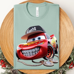 May include: A light green t-shirt featuring a cartoon image of Lightning McQueen with a diamond-studded grill, a backward black cap with the word "Eutts", and a gold chain with diamond pendants. The shirt is on a wooden plate.