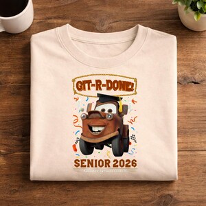 Op de afbeelding: Cr&egrave;mekleurige sweatshirt met een cartoonafbeelding van Takel uit de film Cars, met een afstudeerhoed. De tekst "GIT-R-DONE!" en "SENIOR 2026" staan ook op de trui, samen met confetti en een geblokte vlag.