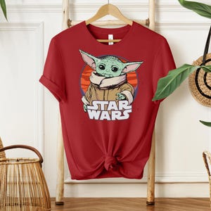 Puede incluir: Camiseta roja con un gr&aacute;fico de un personaje alien&iacute;gena verde con orejas grandes, que lleva un abrigo marr&oacute;n, y el texto "STAR WARS" en blanco. El dise&ntilde;o est&aacute; sobre un fondo circular naranja y rojo.