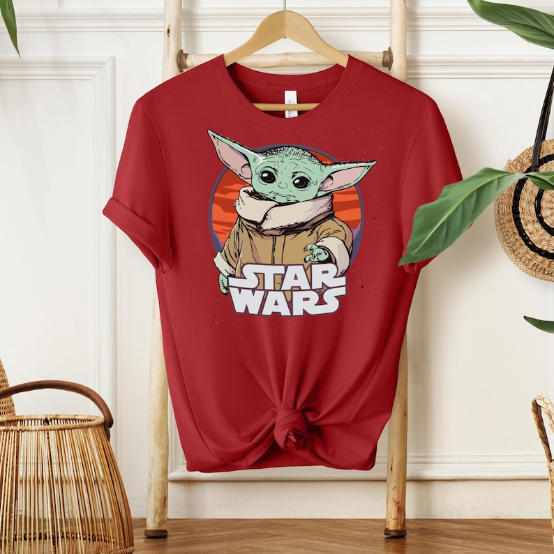 Puede incluir: Camiseta roja con un gr&aacute;fico de un personaje alien&iacute;gena verde con orejas grandes, que lleva un abrigo marr&oacute;n, y el texto "STAR WARS" en blanco. El dise&ntilde;o est&aacute; sobre un fondo circular naranja y rojo.