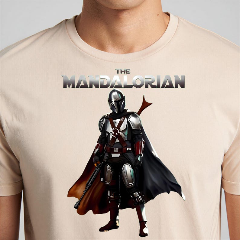 Op de afbeelding: Beige T-shirt met een afbeelding van The Mandalorian, een personage uit de Star Wars-franchise. De afbeelding toont de Mandalorian in volledige uitrusting, met een zwarte cape en een blaster. De tekst "THE MANDALORIAN" staat boven het personage.