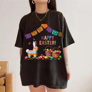 K&ouml;nnte beinhalten: Schwarzes T-Shirt mit einem farbenfrohen "Frohe Ostern!"-Motiv. Das Design umfasst eine Hasen-Pi&ntilde;ata, Osterk&ouml;rbe und dekorative Banner. Schmetterlinge und Ostereier sind ebenfalls abgebildet.