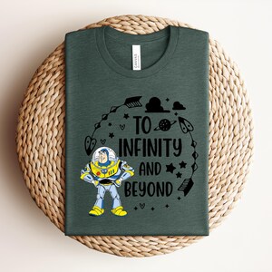 Peut inclure: T-shirt vert fonc&eacute; avec un dessin anim&eacute; de Buzz l'&Eacute;clair et le texte "TO INFINITY AND BEYOND" en noir. Le motif comprend des &eacute;toiles, des fl&egrave;ches et des plan&egrave;tes. Le t-shirt est pr&eacute;sent&eacute; sur une surface tiss&eacute;e.