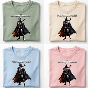 Op de afbeelding: Vier T-shirts in verschillende kleuren: saliegroen, cr&egrave;me, lichtblauw en roze. Elk shirt heeft een afbeelding van het personage The Mandalorian met de tekst "THE MANDALORIAN" boven de afbeelding.
