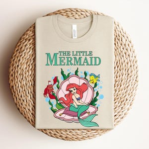 May include: A beige crew neck t-shirt featuring a graphic of The Little Mermaid with Ariel sitting in a pink seashell, accompanied by a red crab and a yellow and blue fish, surrounded by green seaweed and blue bubbles on the front of the shirt.